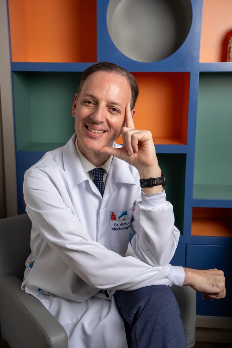 Dr. Victor Horácio sorrindo, sentado diante da estante colorida do consultório