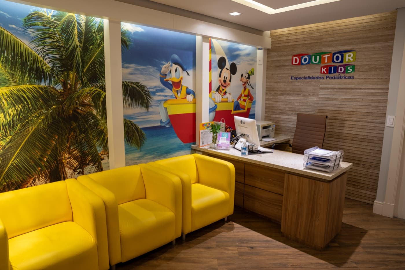 Recepção da Clínica Doutor Kids, com poltronas amarelas e mural do Mickey, Donald e Pateta